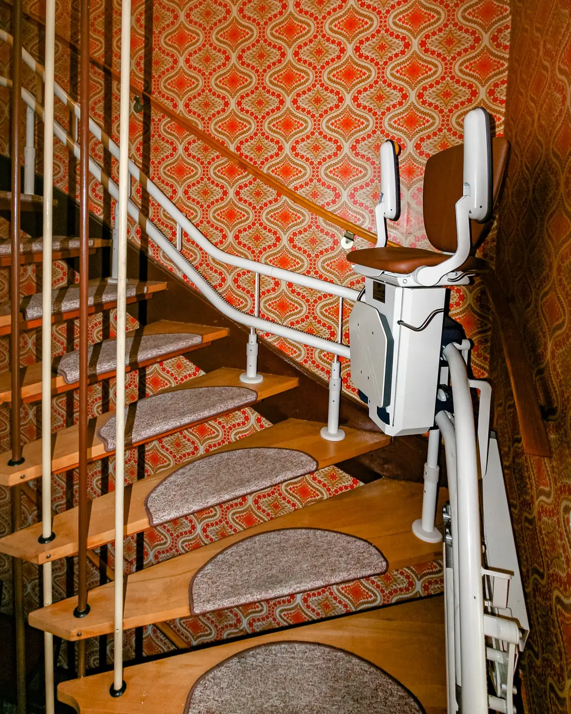 A stairlift fitted to a curved wooden staircase with patterned wallpaper
